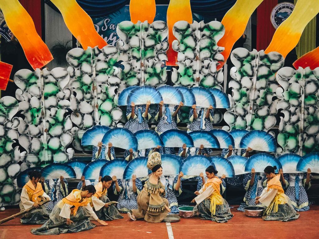 Women in traditional farmer outfits kneeling with water containers during a stage performance at Bugwas Festival, San Juan