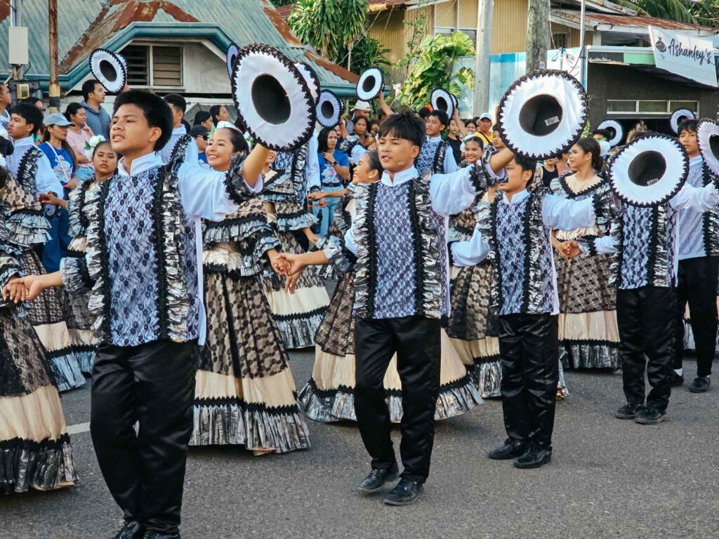 Men and women in black and silver traditional outfits holding hands during a Siquijor street parade, with men proudly raising their hats