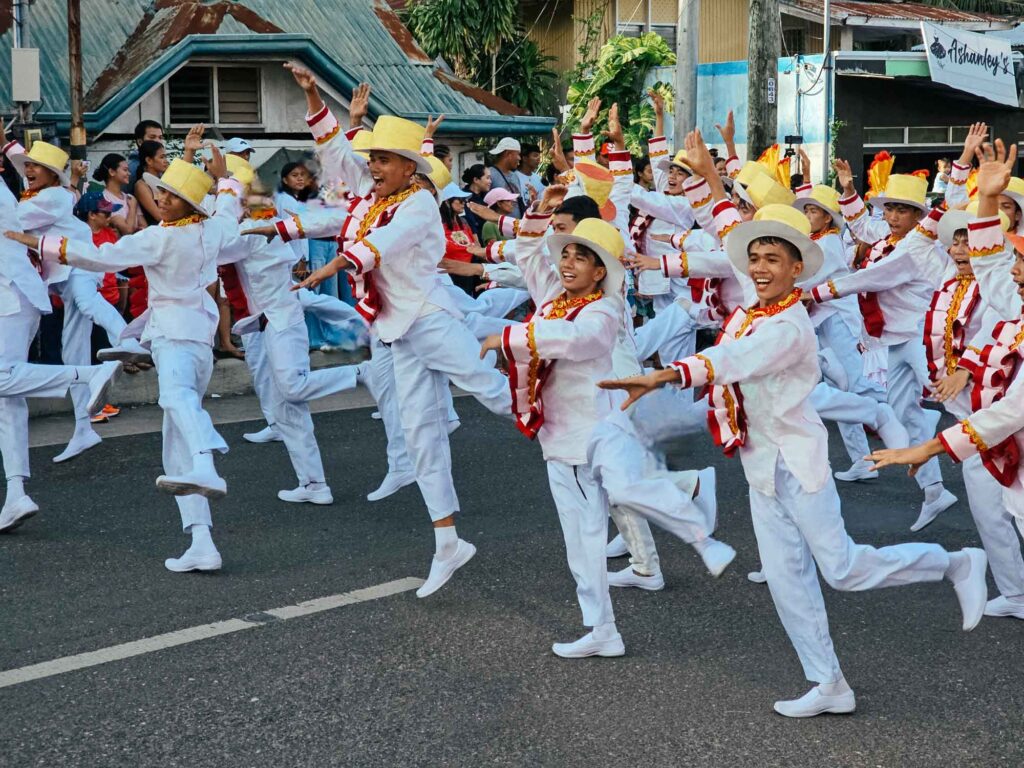 Men in white traditional outfits and yellow hats joyfully jumping during a dance parade at a Siquijor fiesta